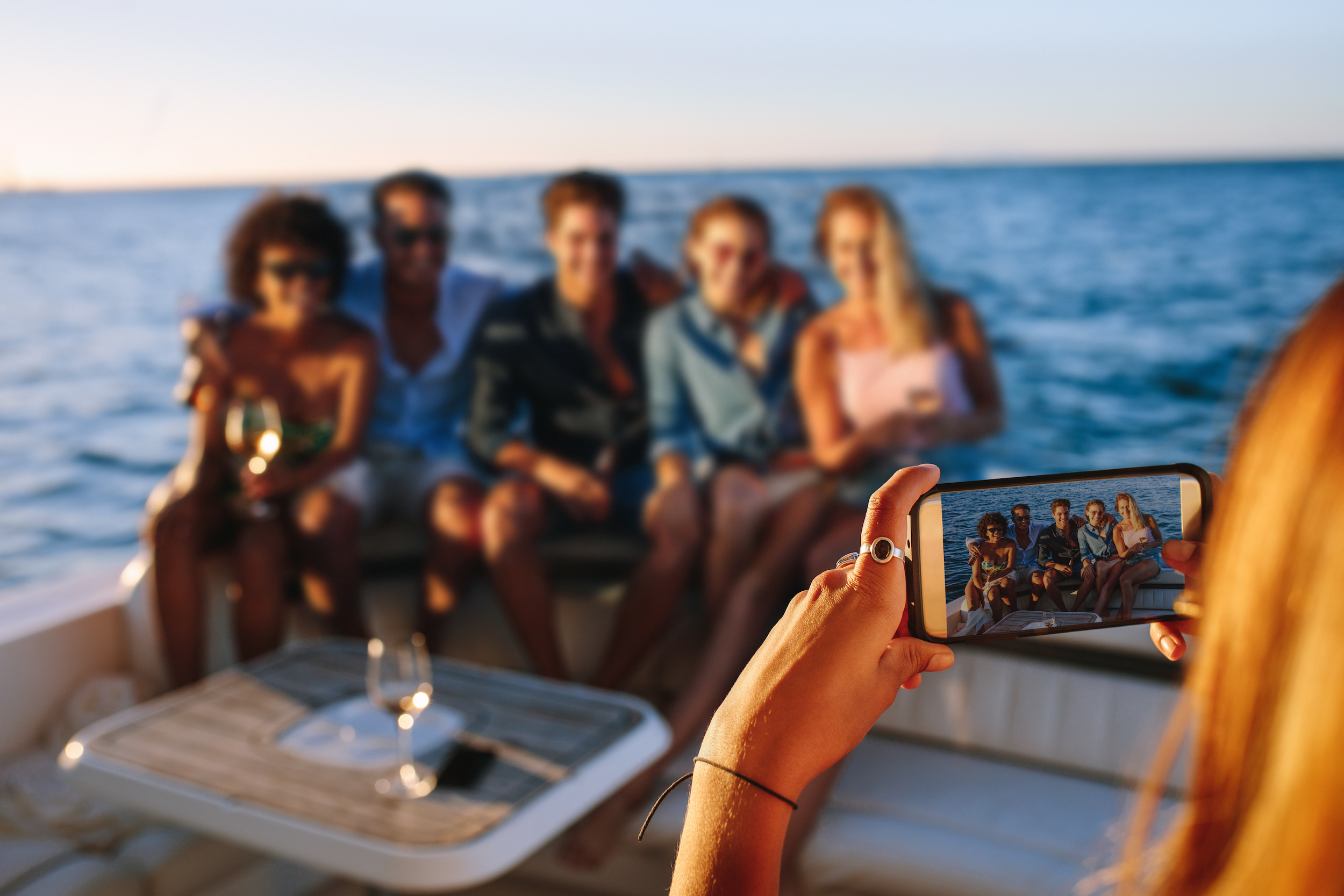 Woman taking a picture with mobile phone of her friends sitting on boat. Capturing memories of boat party.